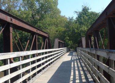 Sunny paved trail with wood rails and a bridge going over water